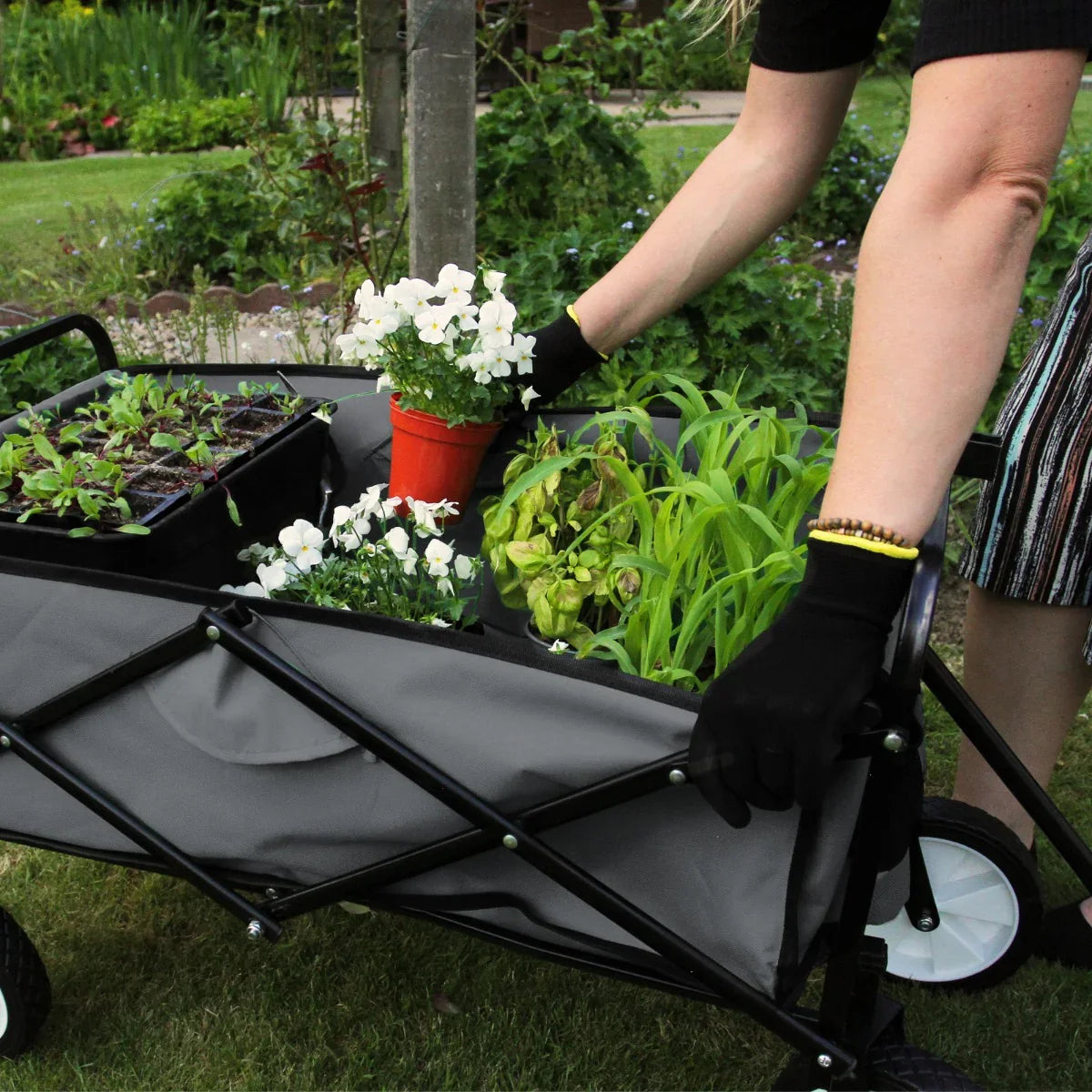 Foldable grey garden cart with black handles, filled with potted plants and flowers, being pushed on a grassy area