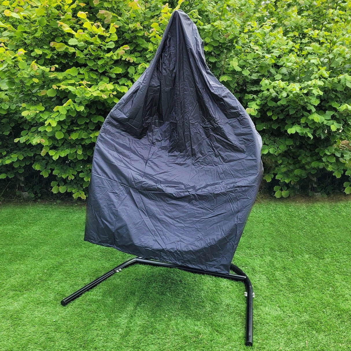 Black hanging rattan egg chair covered with a protective tarp, set against a lush green hedge on a grassy area