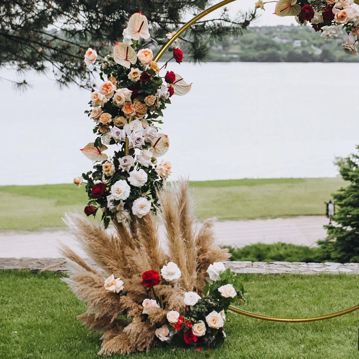 Gold wedding moongate arch adorned with white and red roses, lush greenery, and pampas grass, set against a serene waterfront backdrop