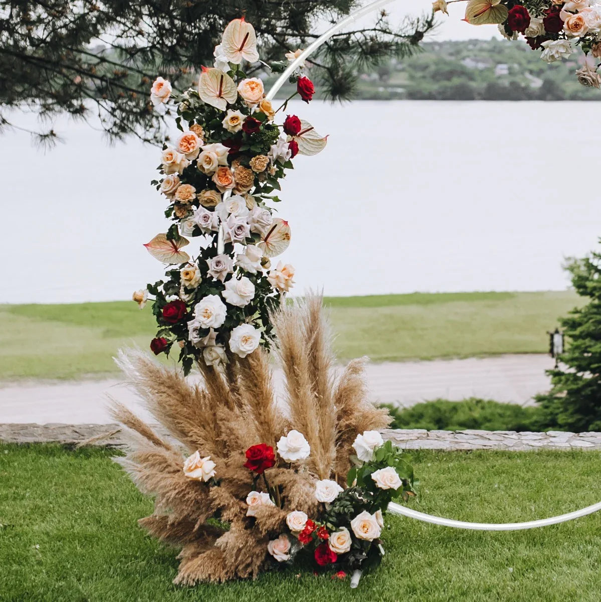 Elegant white wedding moongate adorned with lush roses and pampas grass, set against a serene outdoor backdrop by the water