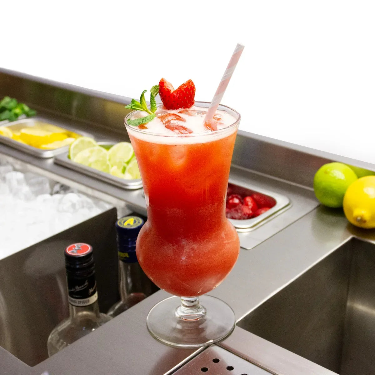 Tall glass filled with vibrant red cocktail, garnished with strawberries and mint, set on a sleek stainless steel bar station