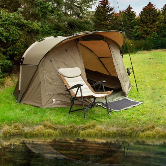 Spacious beige camping tent set up on green grass near a calm pond, with a folding chair positioned outside for relaxation