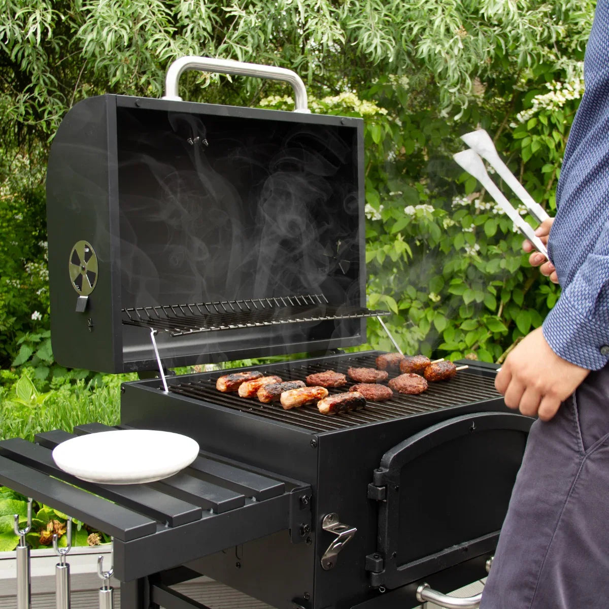 XL BBQ smoker with a black finish, grilling sausages and burgers, surrounded by greenery and emitting smoke, showcasing its spacious cooking area
