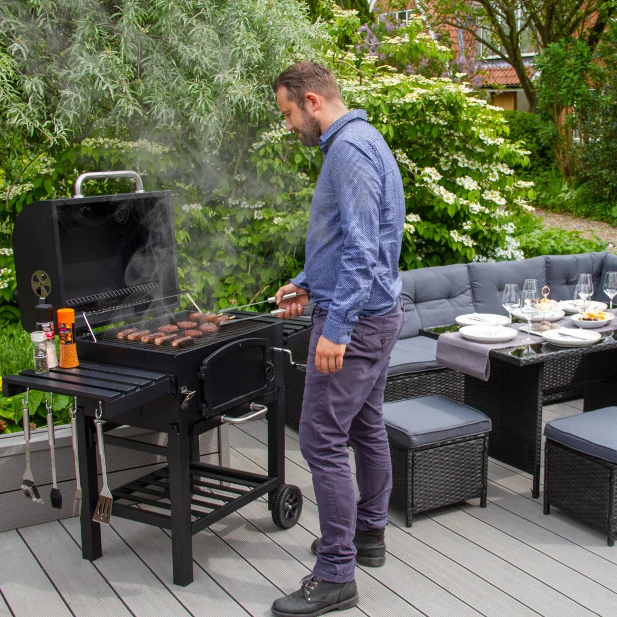 XL BBQ smoker with sausages grilling, man in blue shirt, outdoor patio setting with greenery and dining setup in background