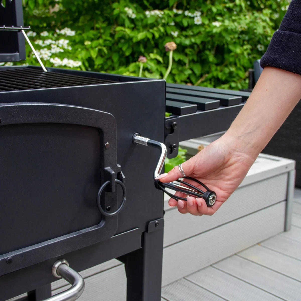 XL BBQ smoker with a black metal finish, featuring a hand adjusting the side crank for temperature control amidst greenery
