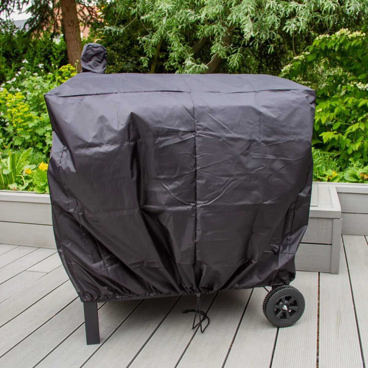 XXL BBQ smoker covered with a durable black protective cover, featuring wheels for easy mobility, set on a wooden deck surrounded by greenery
