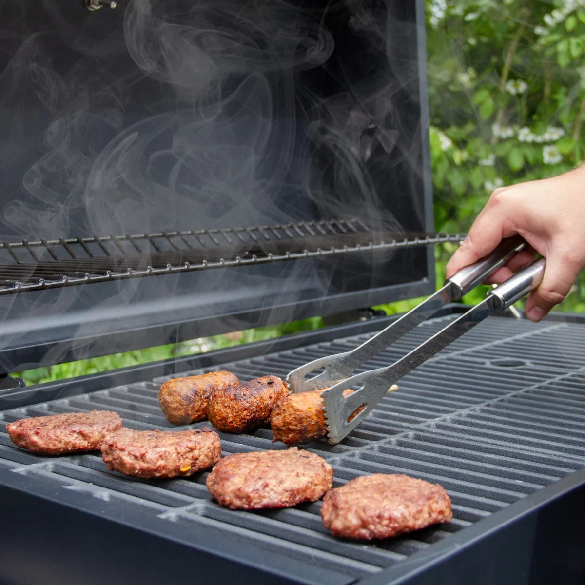 XXL BBQ smoker with smoky grilled burgers on the cooking grate, showcasing stainless steel tongs and a lush green background