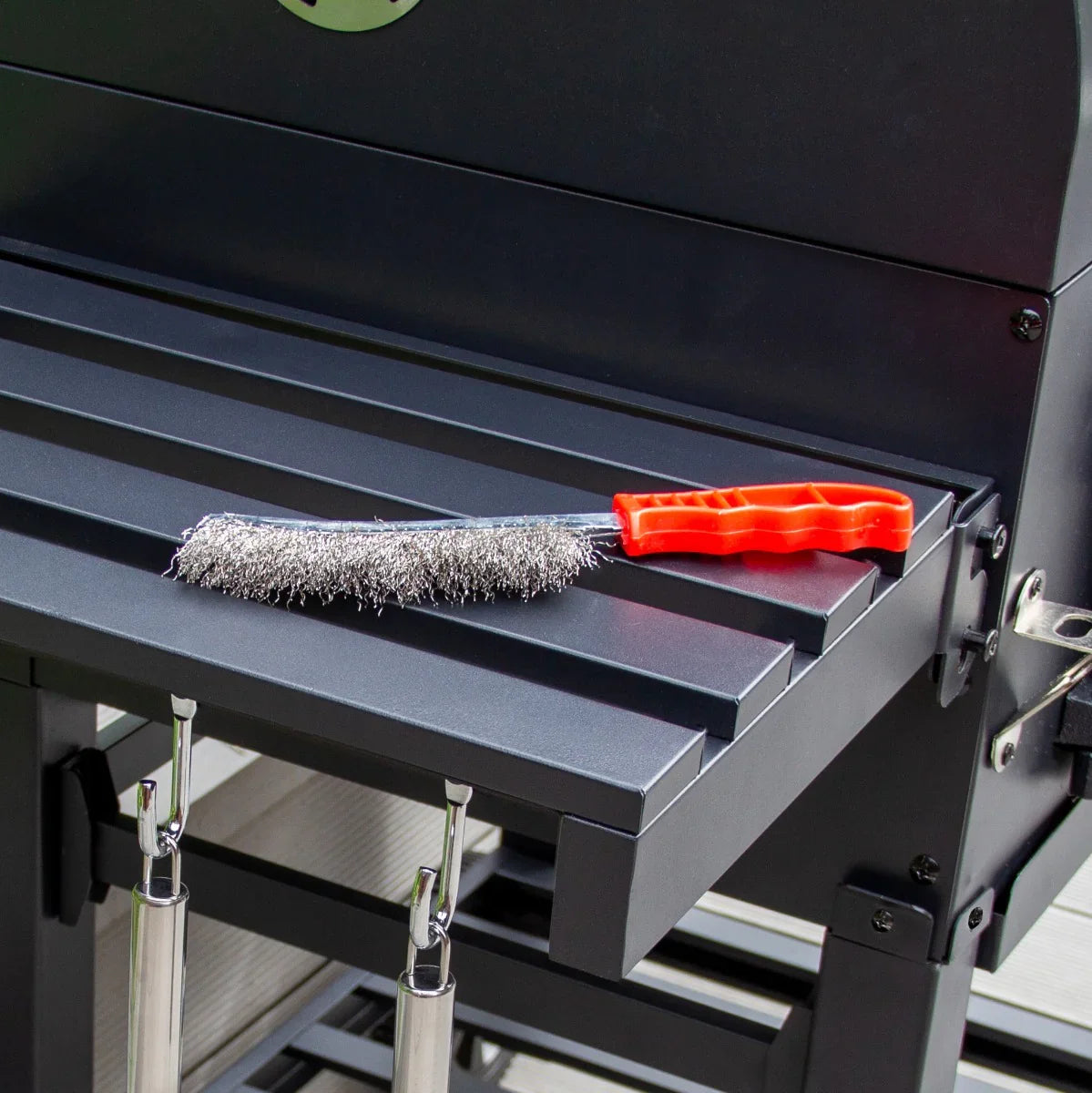 Stainless steel grill brush with a red handle resting on the black metal surface of the XXL BBQ smoker, ideal for cleaning grates