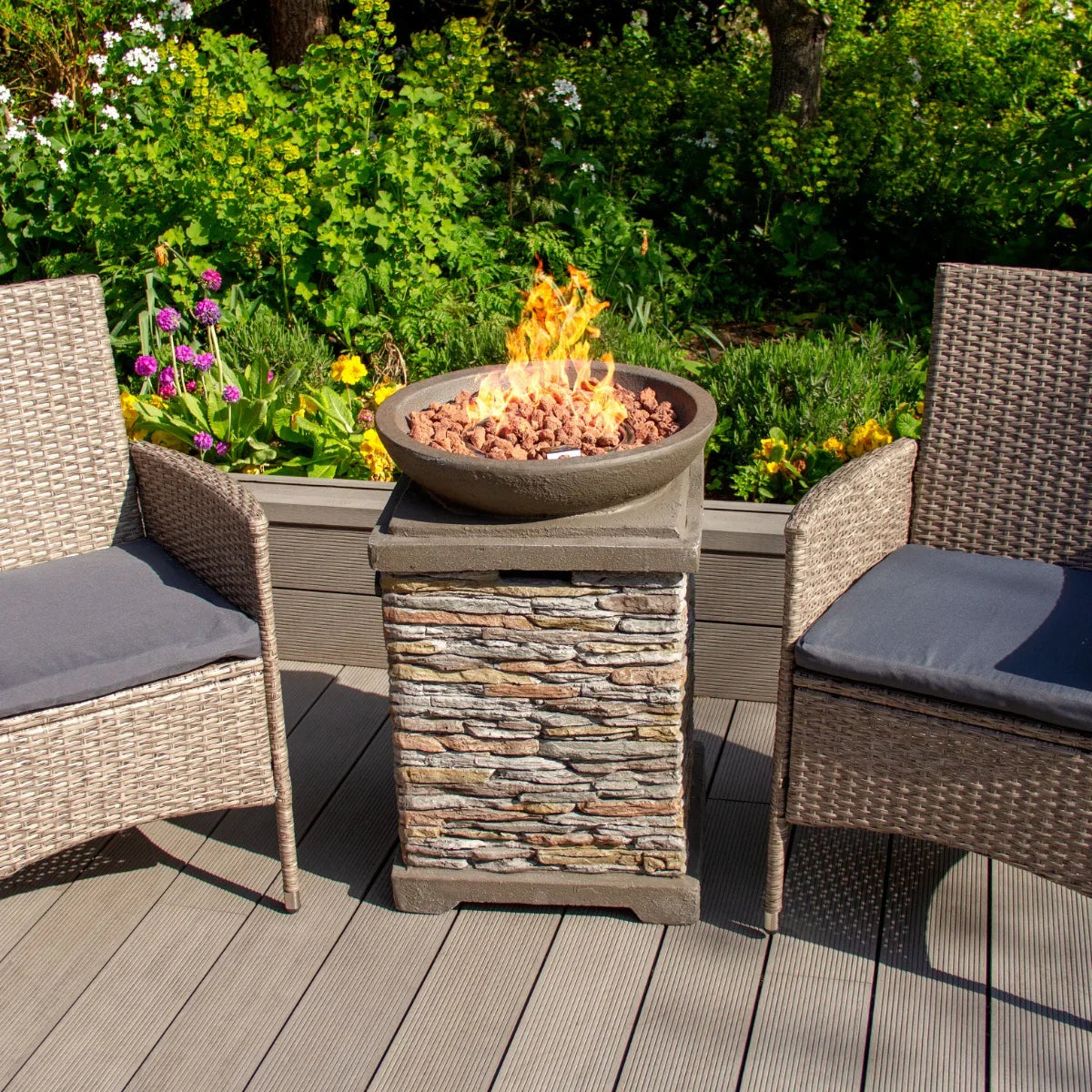 Stone gas fire pit heater with a round bowl, textured stone base, surrounded by two wicker chairs and vibrant garden flowers