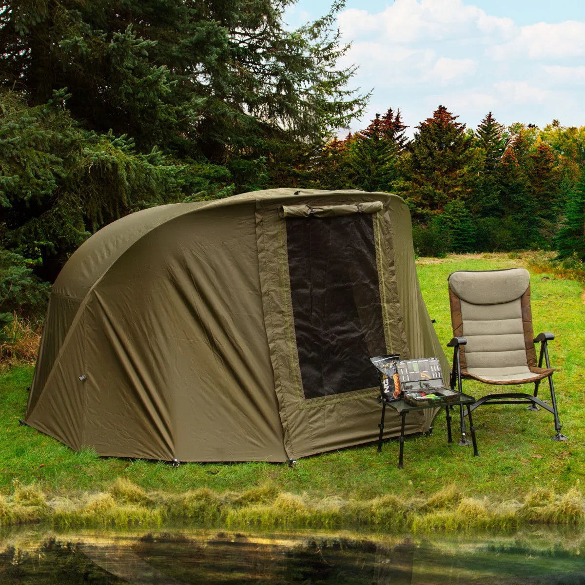 Two-man fishing bivvy in olive green with a mesh window, set beside a folding chair and tackle box near a serene lake