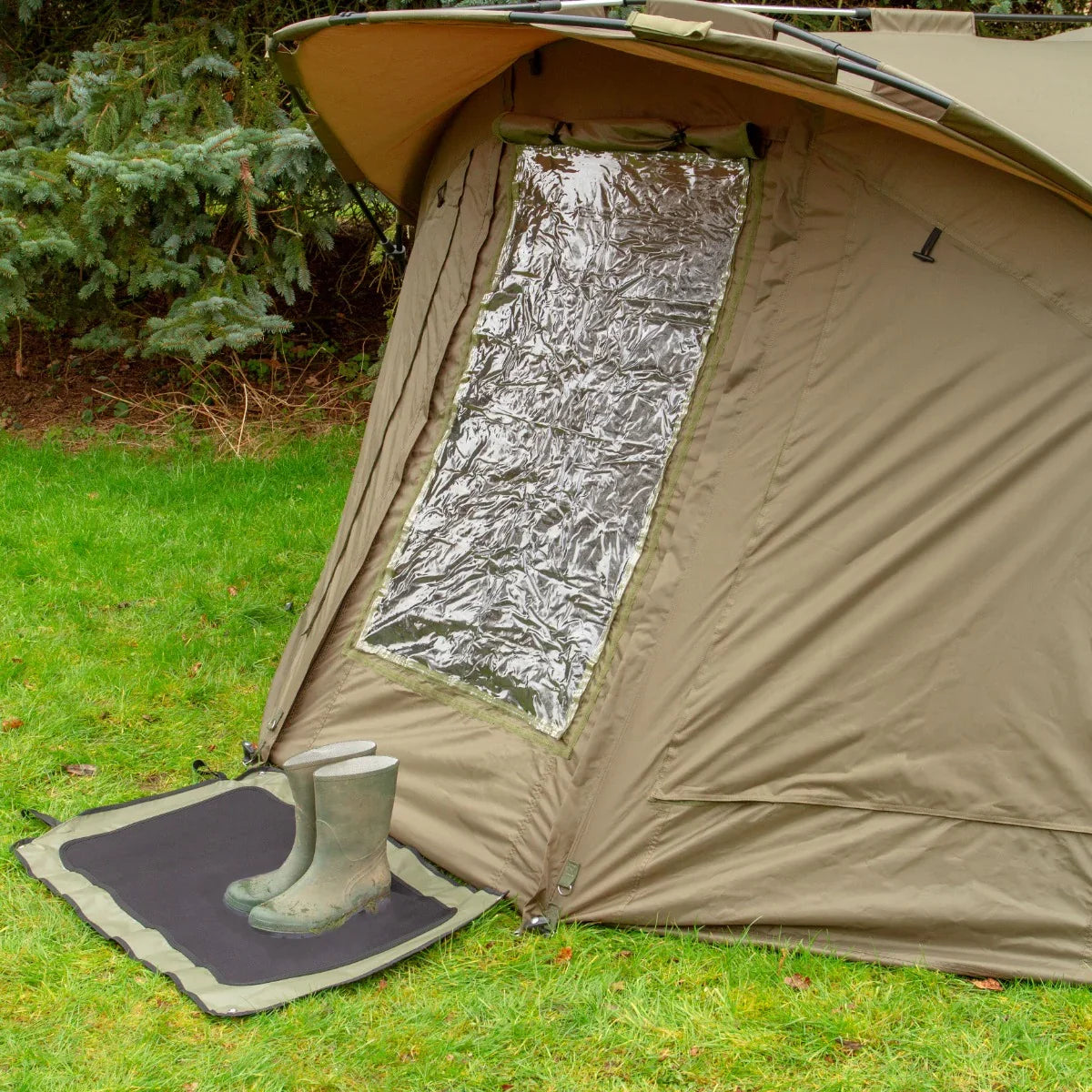 Two-man fishing bivvy in khaki with a large transparent window, set on grass, featuring a boot mat and rubber boots nearby