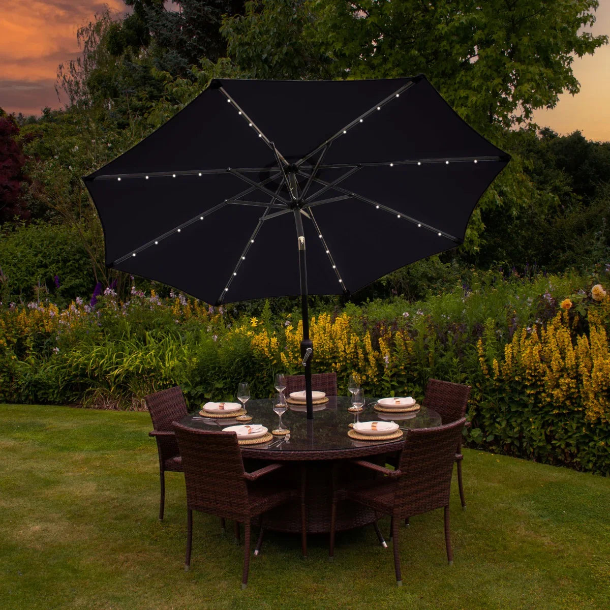 Black 2.7m LED tilt parasol with string lights, providing shade over a round dining table set in a vibrant garden setting