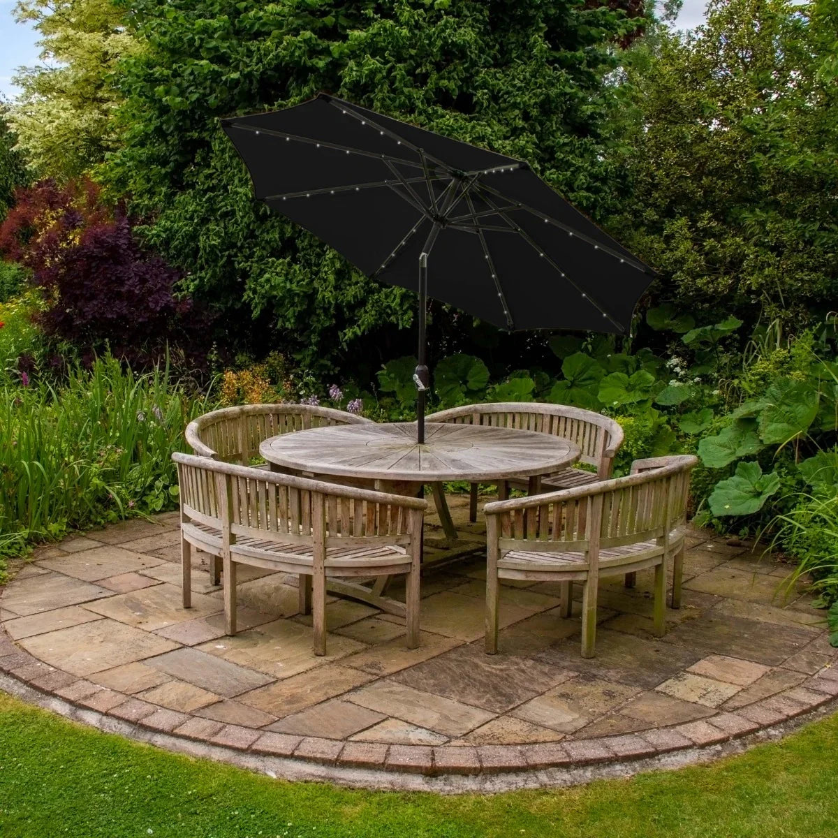 Black 2.7m LED tilt parasol providing shade over a round wooden garden table surrounded by four chairs in a lush green setting