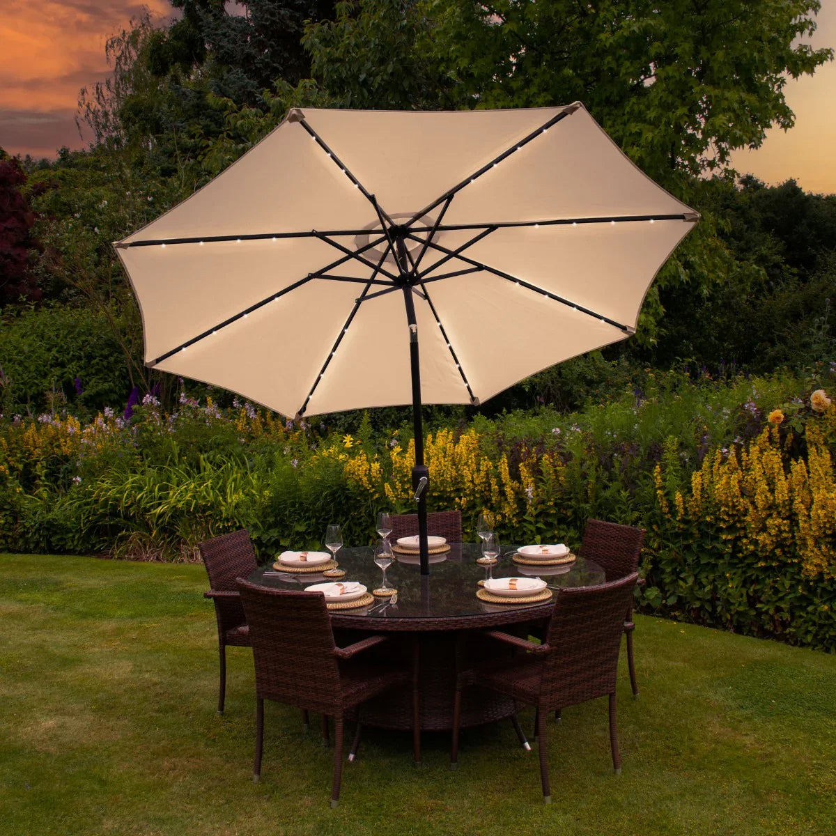 Beige 2.7m LED tilt parasol with a sturdy black frame, illuminating a dining table set in a lush garden with colorful flowers