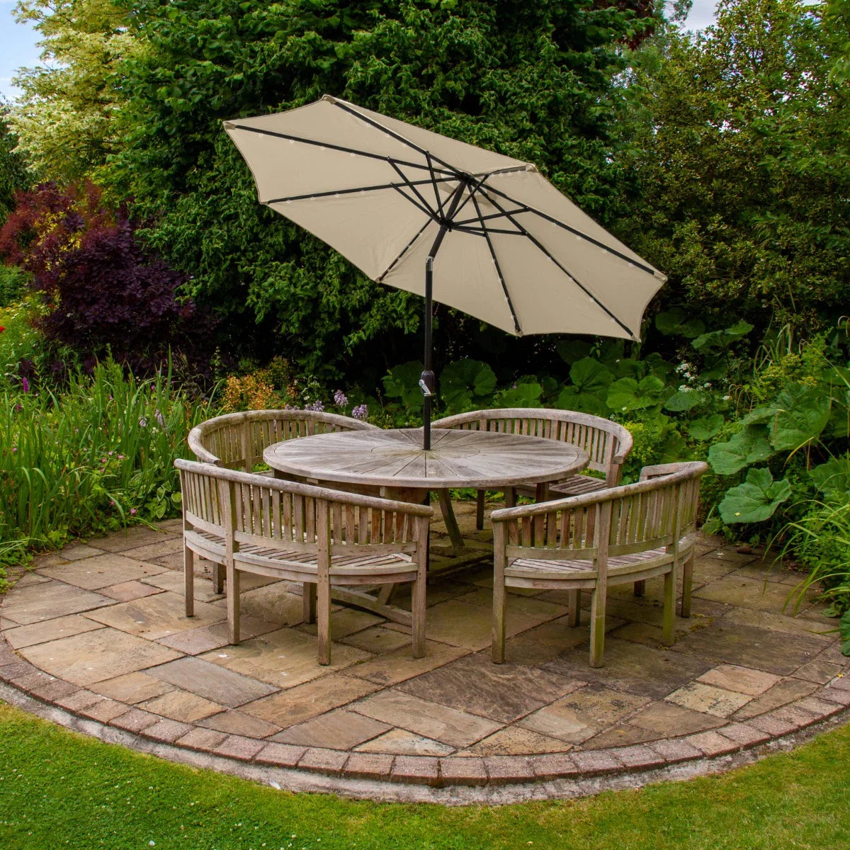 Beige 2.7m LED tilt parasol positioned over a wooden circular table surrounded by four chairs in a lush garden setting