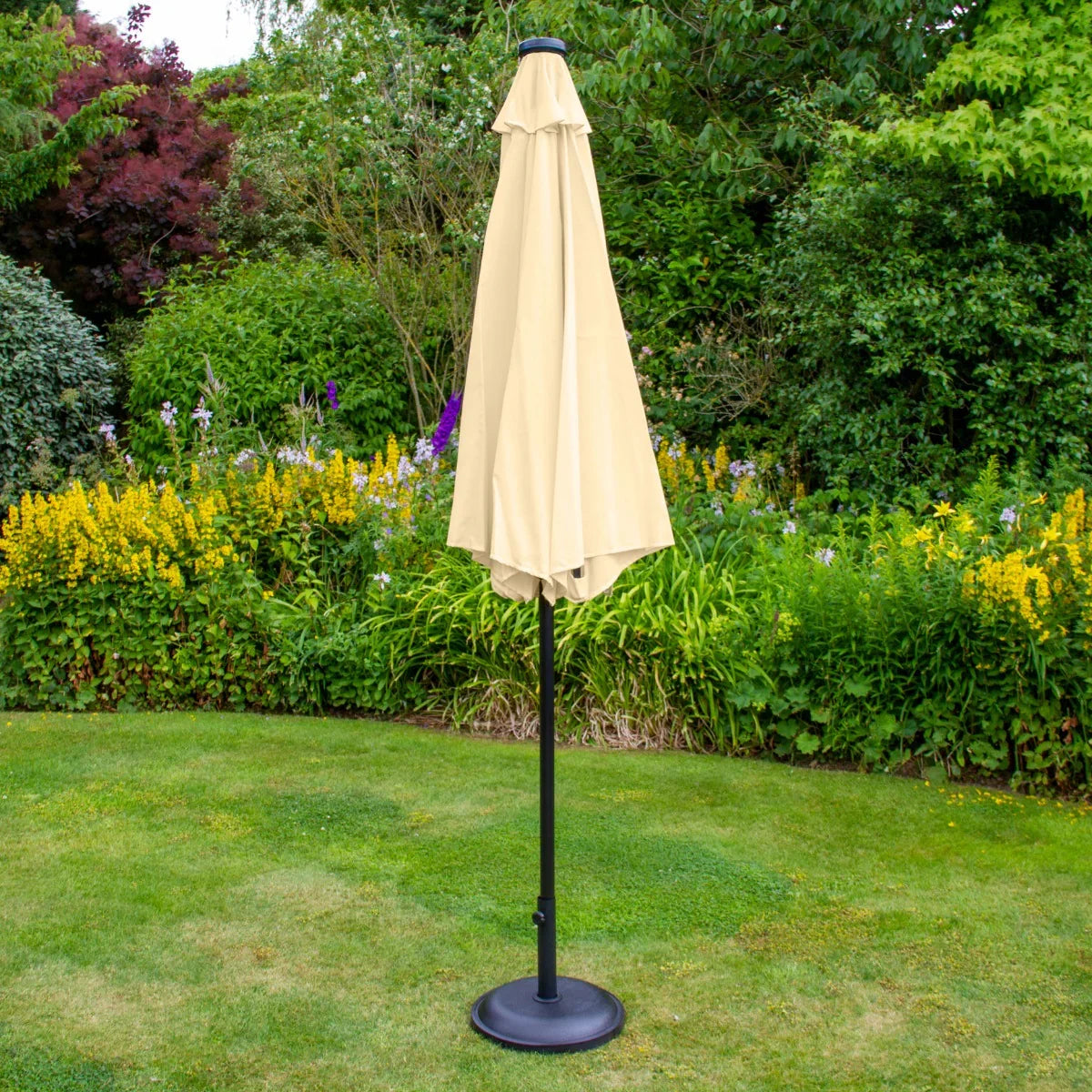 Beige 2.7m LED tilt parasol with a sleek black pole, standing in a garden surrounded by vibrant flowers and greenery