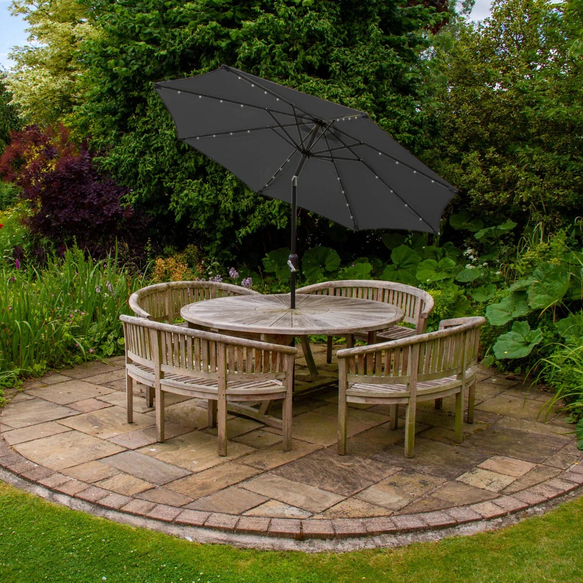 Grey 2.7m LED tilt parasol with a sleek design, positioned above a round wooden table surrounded by rustic chairs in a lush garden setting