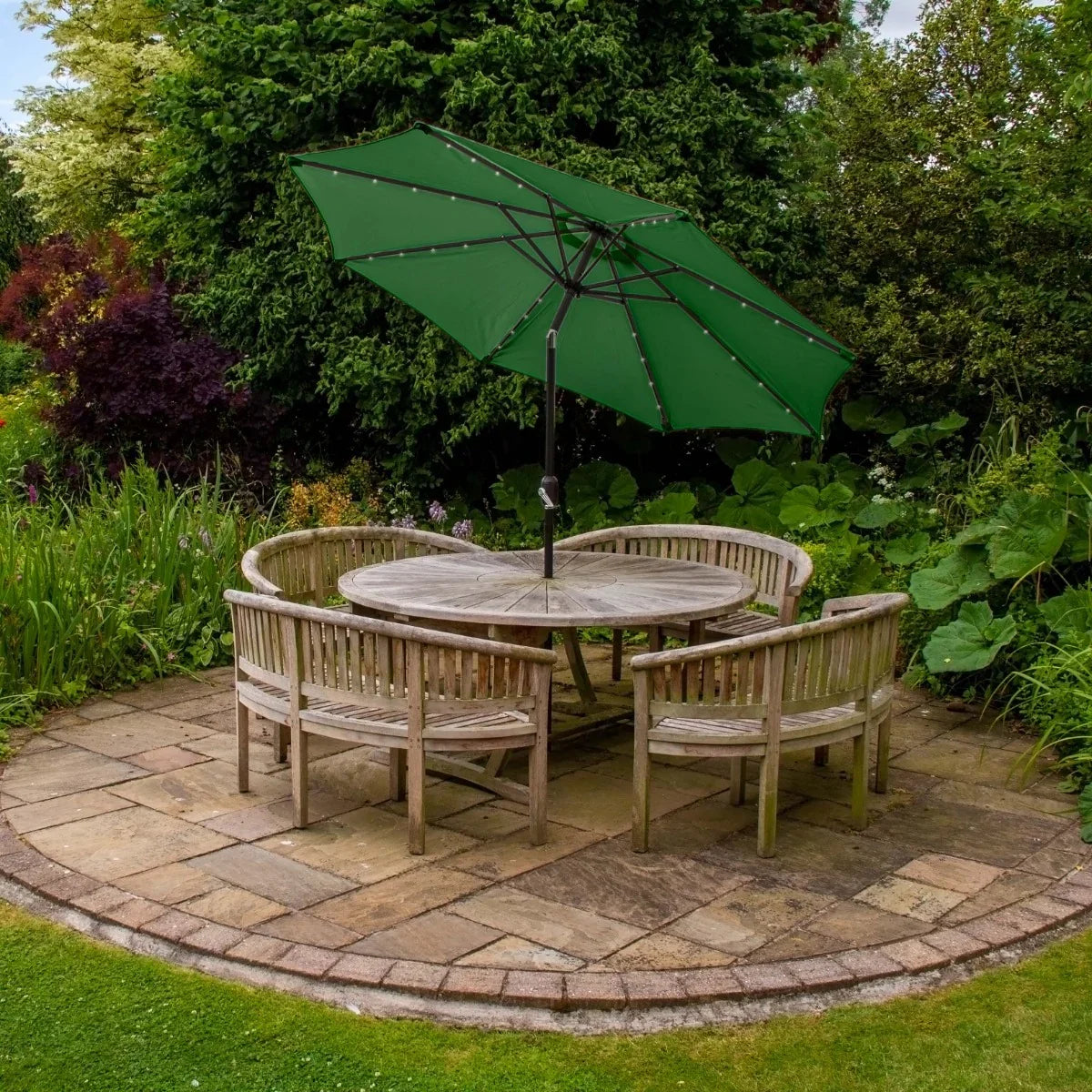 Green 2.7m LED tilt parasol positioned over a circular wooden table surrounded by rustic chairs in a lush garden setting