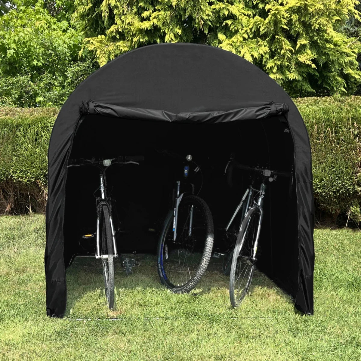 Large black bike tent with a rounded top, sheltering two bicycles, set against a lush green background of shrubs and grass