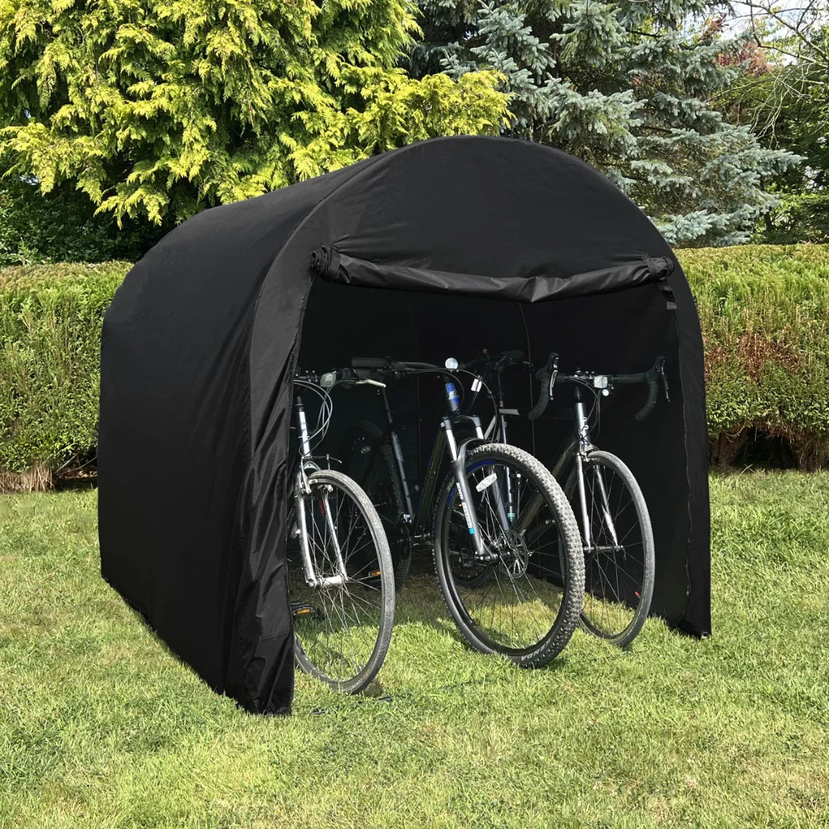 Large black bike tent with a rounded top, sheltering three bicycles, set against a green hedge and trees in a sunny outdoor setting