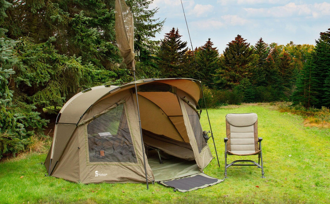 Spacious olive green camping tent with mesh windows, set on grassy ground beside a reclining chair and surrounded by pine trees