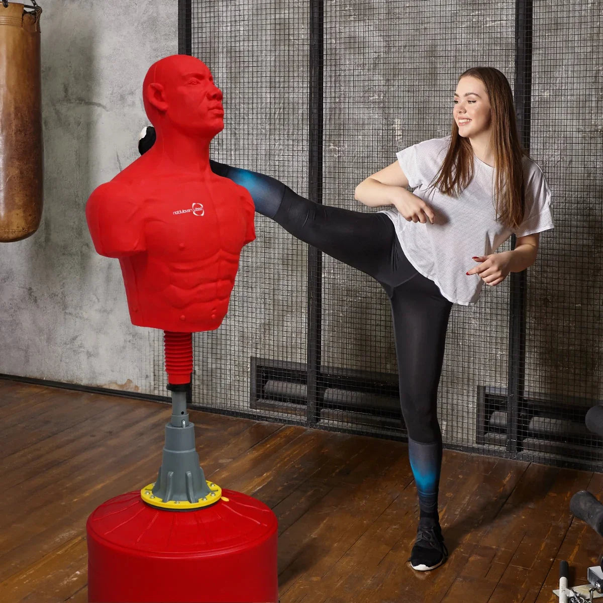 Large free-standing boxing dummy in red, featuring a muscular torso and adjustable height, with a person delivering a kick in a gym setting
