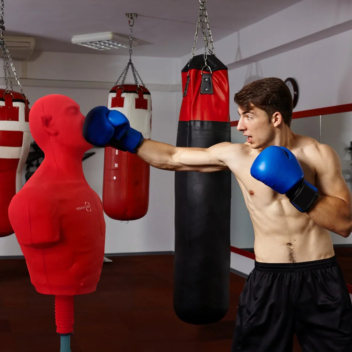 Large free-standing boxing dummy in red, designed for training, with a muscular shape, used by a boxer wearing blue gloves in a gym setting