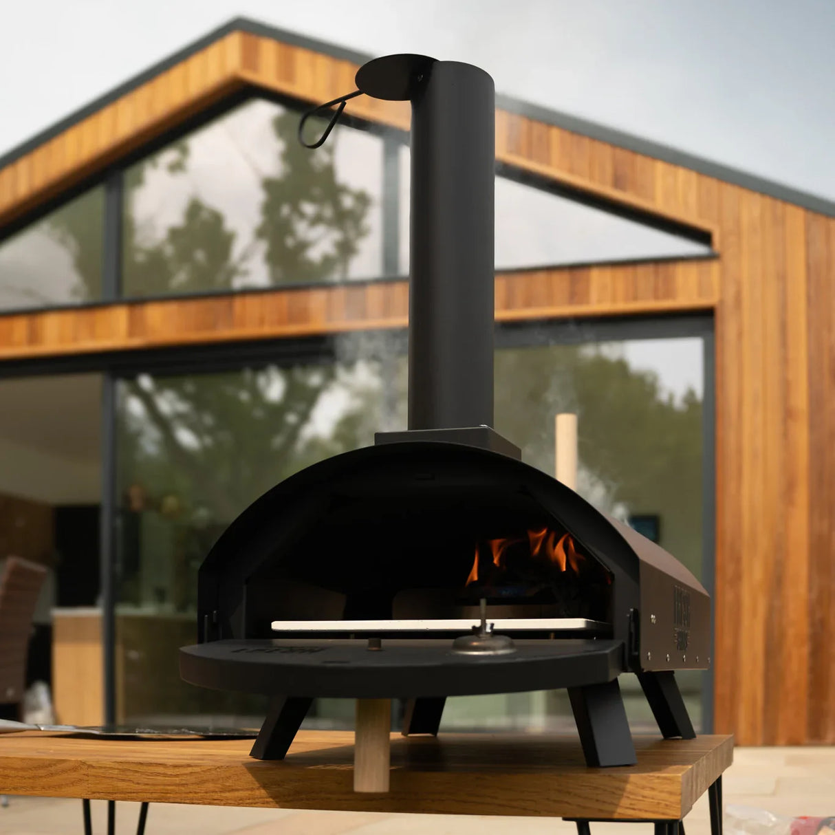 Classic outdoor pizza oven with a sleek black finish, chimney, and flames visible inside, set against a modern wooden backdrop