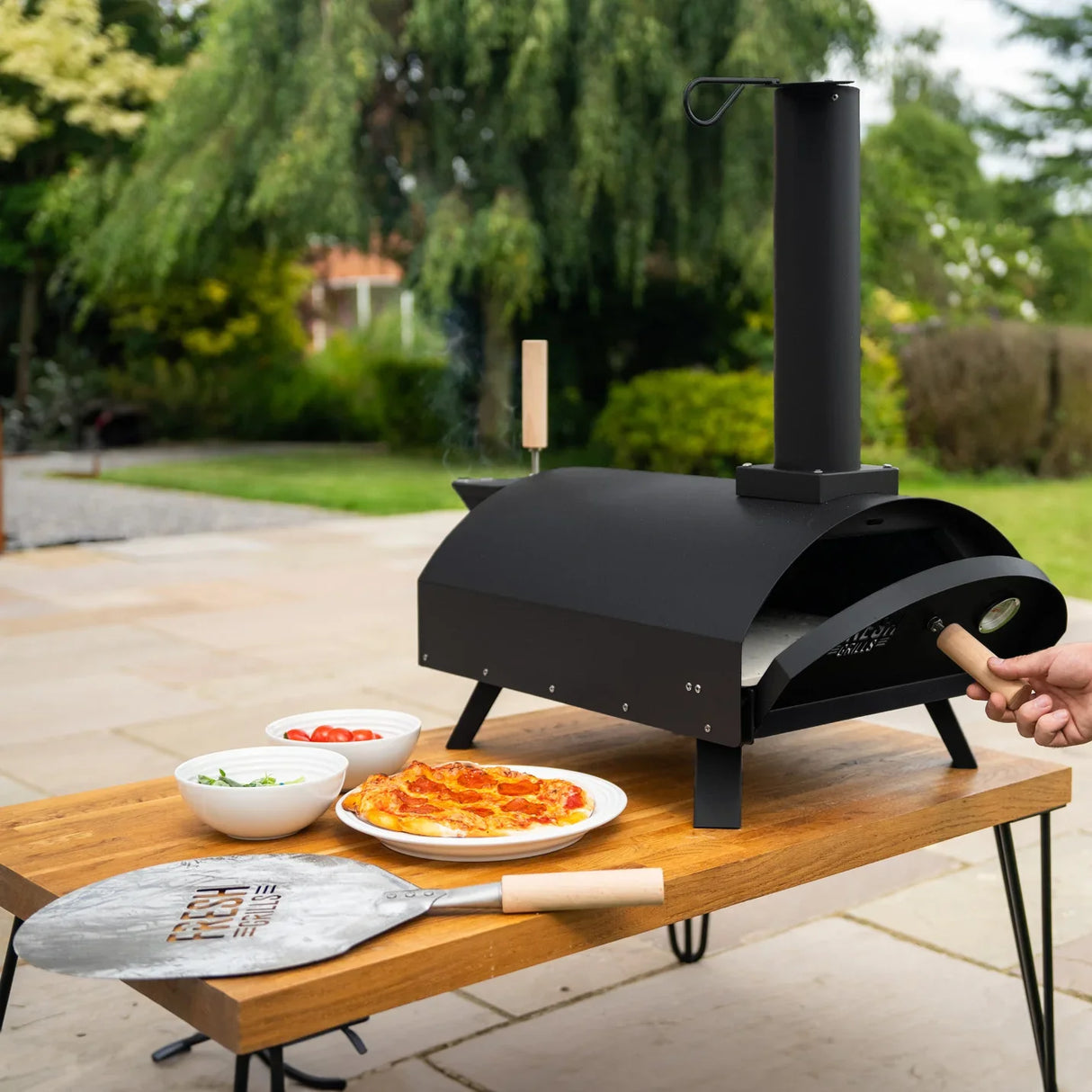 Classic outdoor pizza oven in sleek black with chimney, placed on a wooden table alongside pizza and fresh toppings