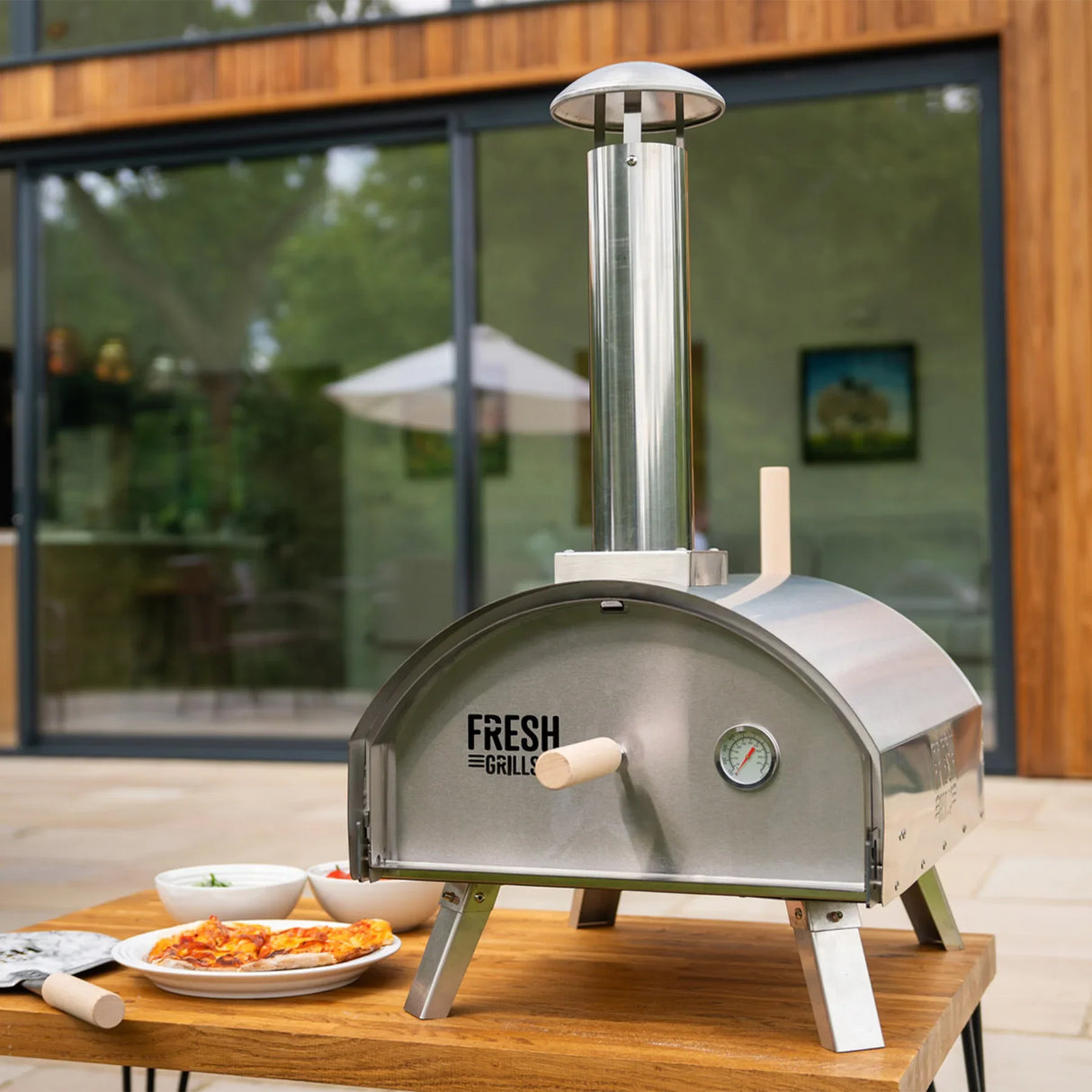 Premium stainless steel outdoor pizza oven with a chimney, temperature gauge, and wooden handles, set on a wooden table outdoors