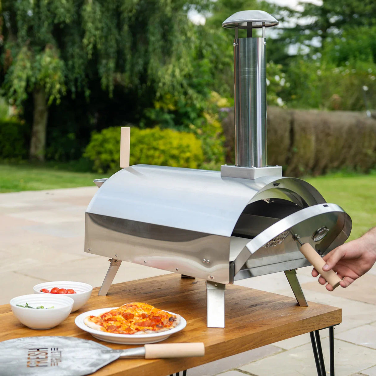 Premium stainless steel outdoor pizza oven with a chimney, set on a wooden table alongside fresh pizzas and toppings