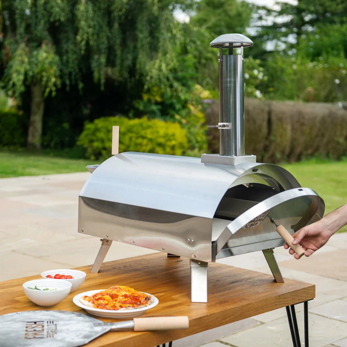 Extra large premium outdoor pizza oven in stainless steel with a chimney, set on a wooden table with fresh ingredients and pizza