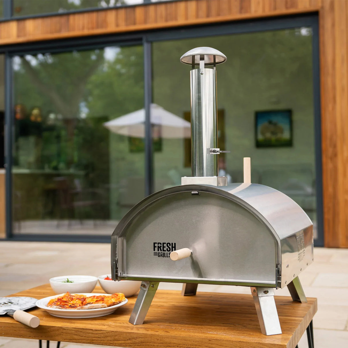 Extra large premium outdoor pizza oven in stainless steel with a chimney, placed on a wooden table with pizza and toppings nearby