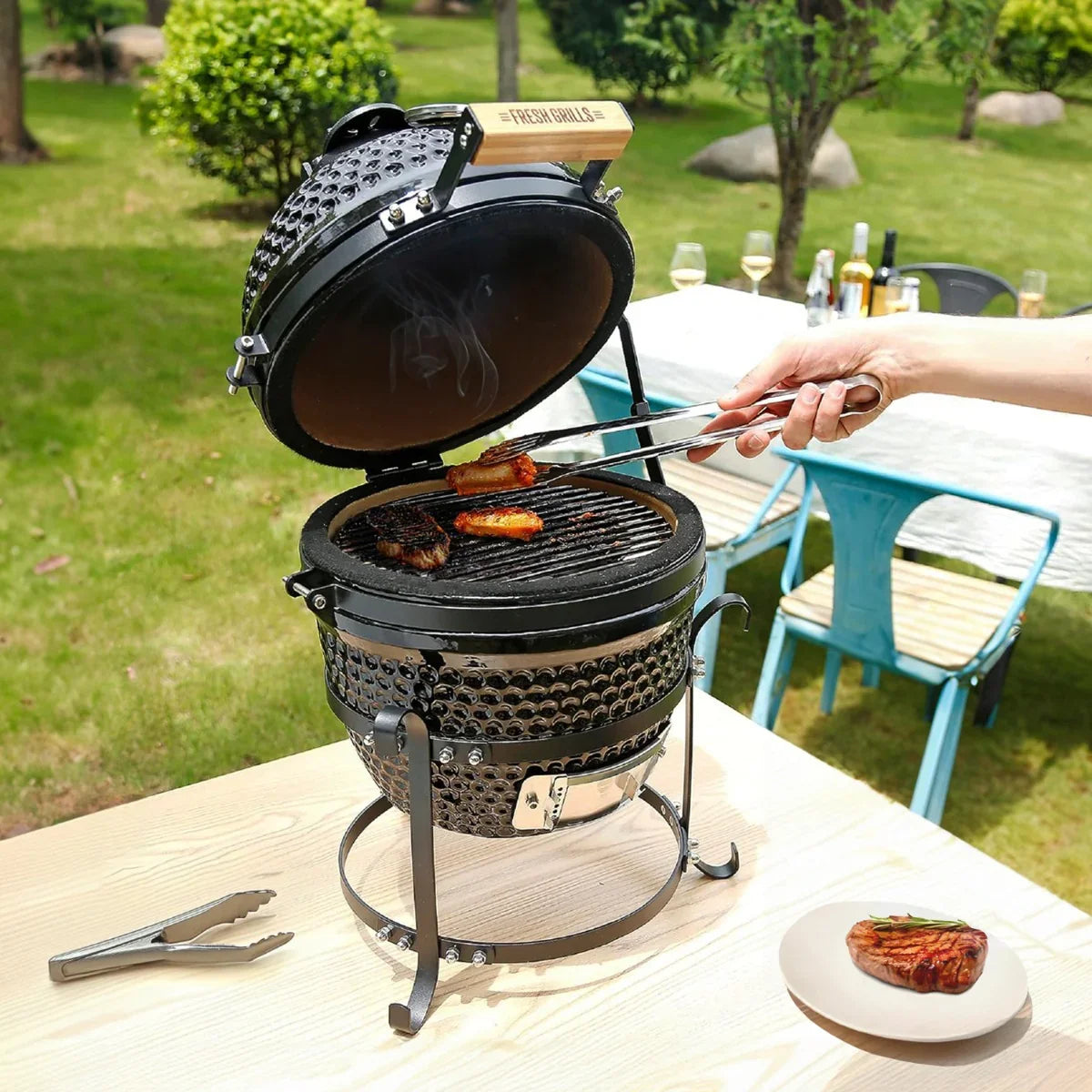 Fresh Grills 13" Kamado BBQ Oven with a glossy black finish, grilling steaks on a charcoal grill, set outdoors on a wooden table