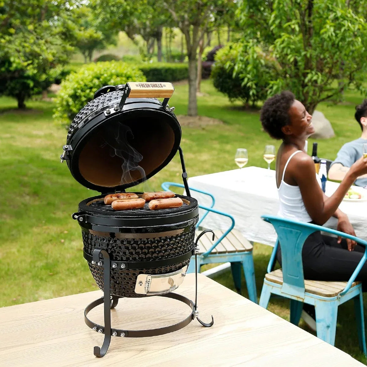 Fresh Grills 13" Kamado BBQ Oven with a glossy black finish, grilling sausages, set in a lush outdoor dining area with guests