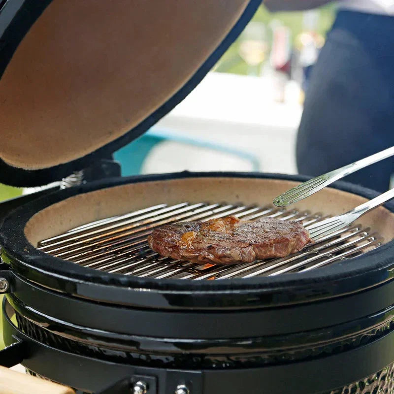 Kamado charcoal BBQ grill with a glossy black finish, featuring a spacious cooking grate and a perfectly seared steak on the grill