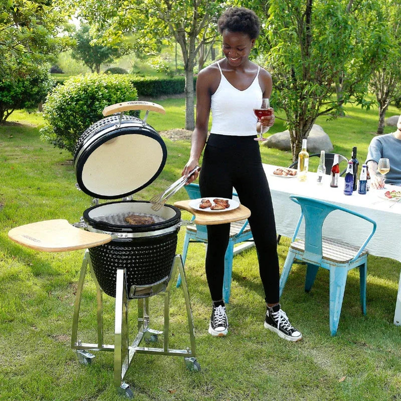 Fresh Grills Kamado Charcoal BBQ Grill 21" in use outdoors, featuring a black ceramic finish, wooden side tables, and a woman grilling food