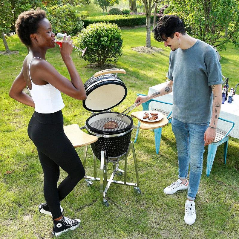 Kamado charcoal BBQ grill in black with a glossy finish, surrounded by people enjoying a sunny outdoor barbecue setting
