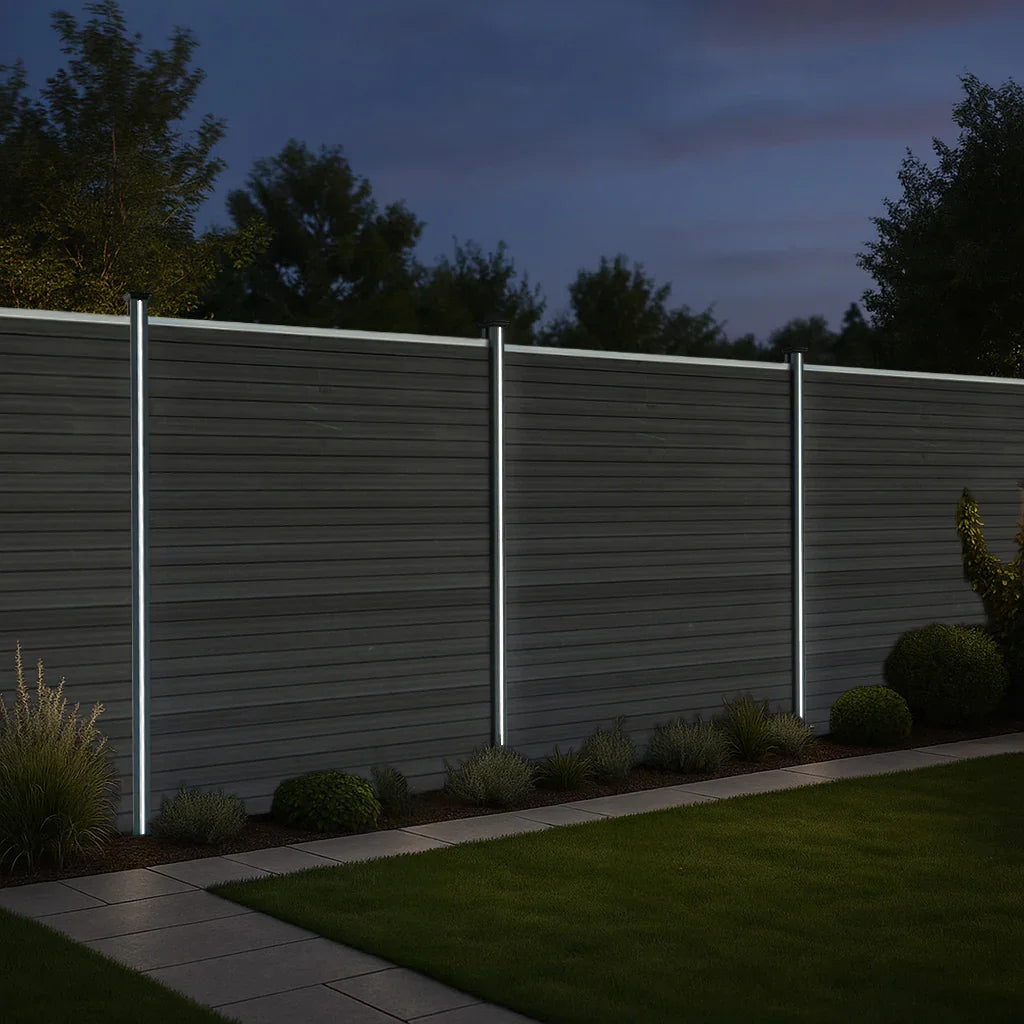 Grey composite fence panels with sleek LED posts, illuminated at night, surrounded by lush greenery and a stone pathway