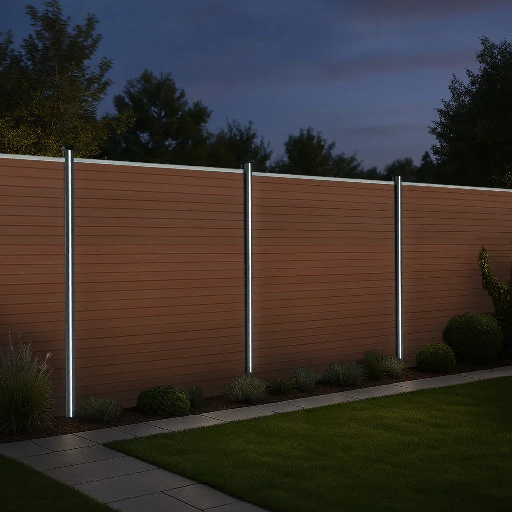 Brown composite fence panels with angled pieces and LED posts, illuminated at night, showcasing a sleek modern design in a garden setting