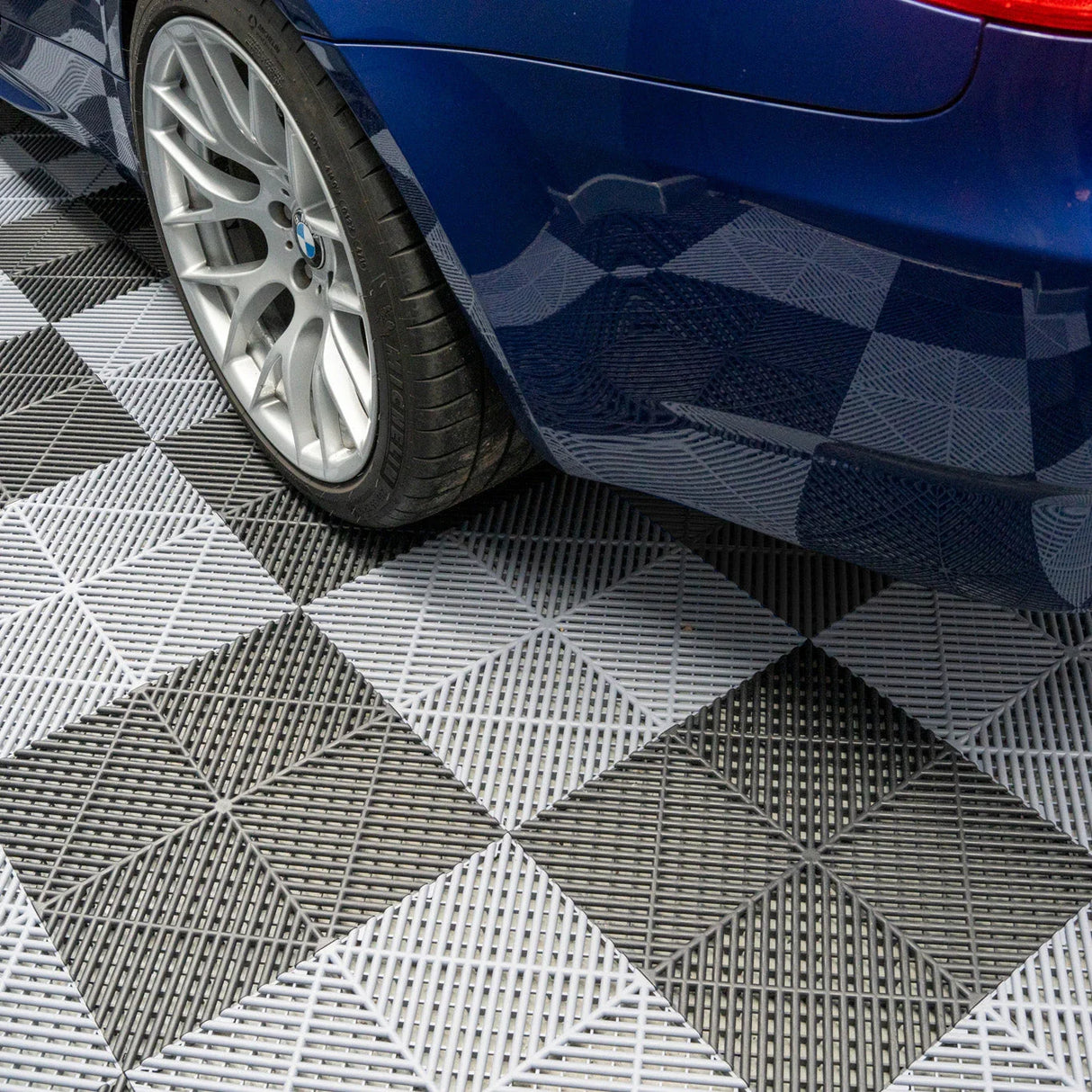 Vented garage floor tiles in black and grey, featuring a checkered pattern, support a sleek blue car with alloy wheels
