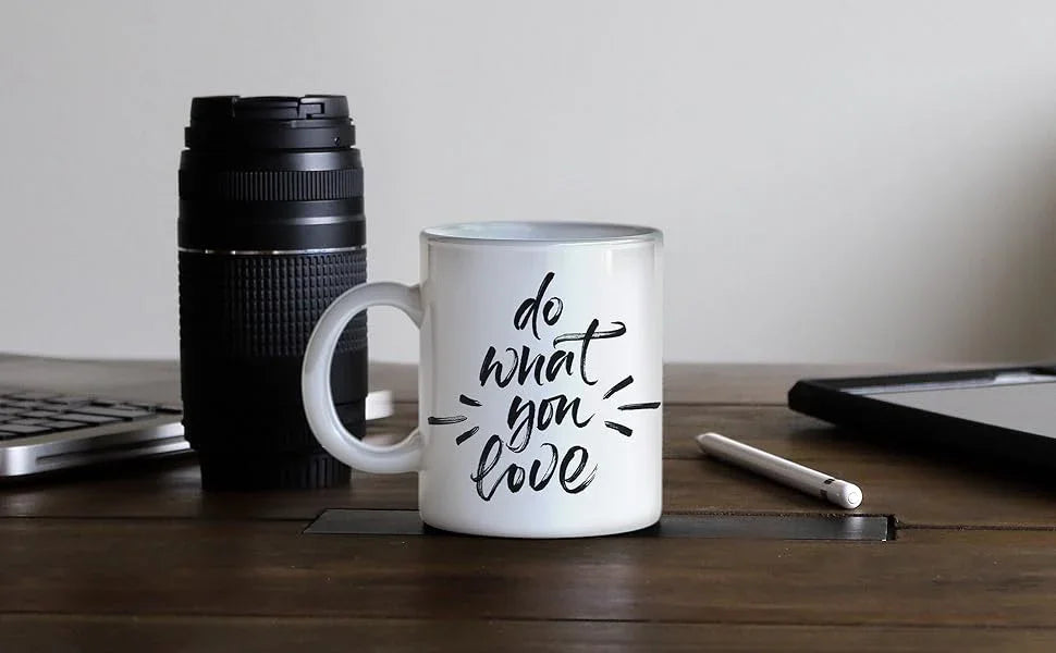 White ceramic mug with black handwritten text "do what you love," placed on a wooden table beside a camera lens and tablet