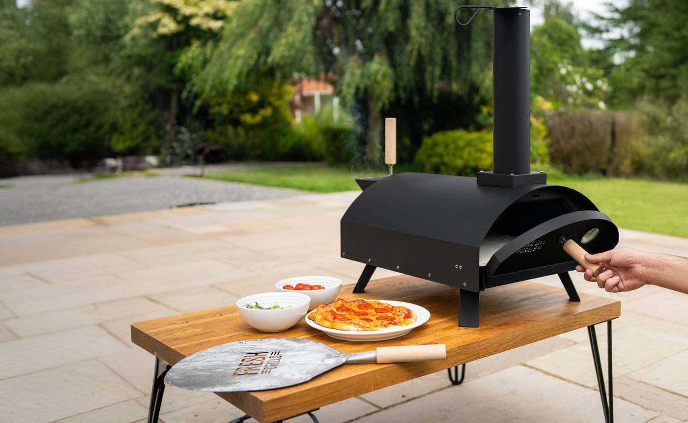 Outdoor black pizza oven on a wooden table, with fresh pizza and toppings, set in a lush garden environment
