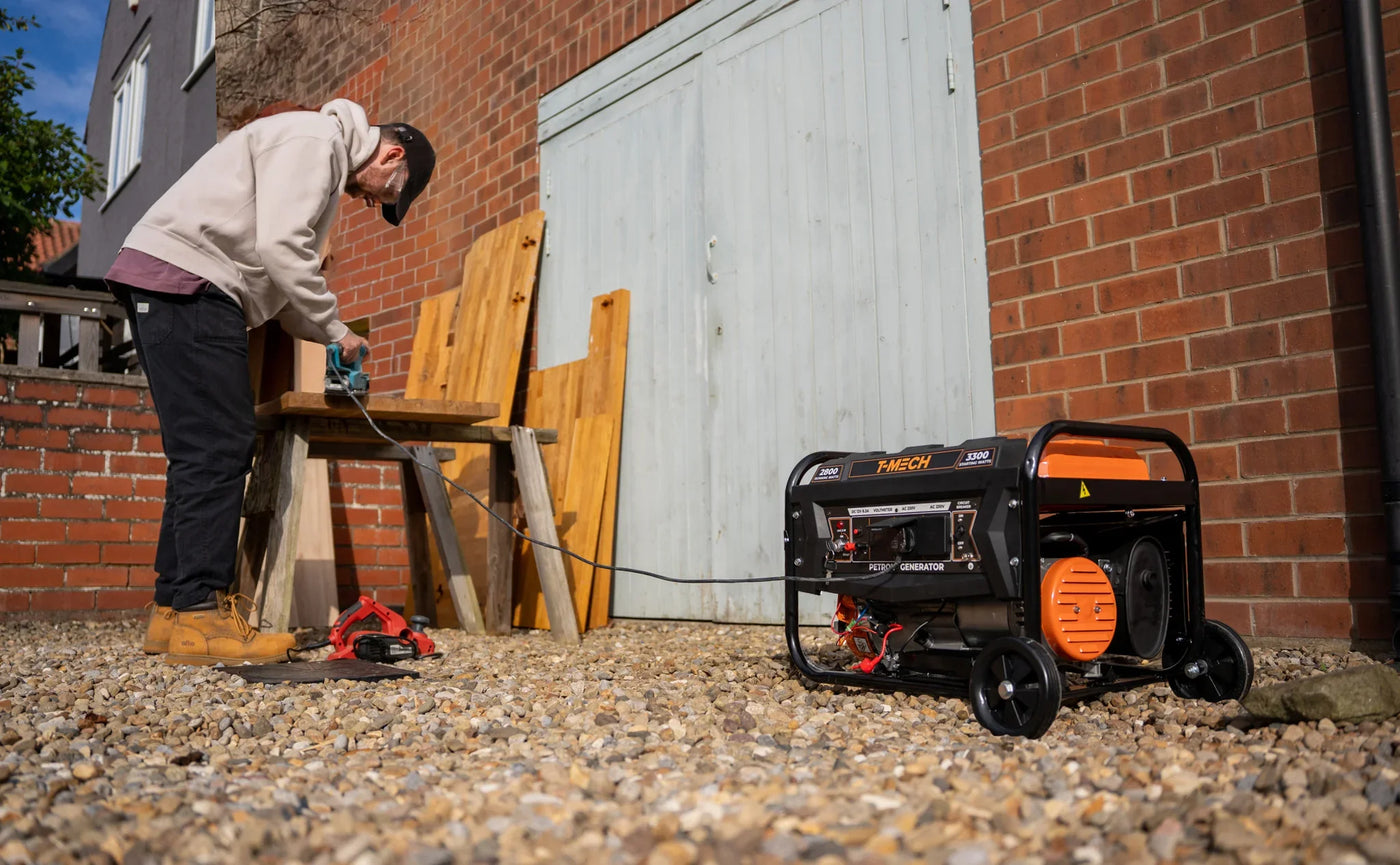 A man in a hoodie connects a power tool to a portable generator on a gravel surface near a brick wall and wooden bench