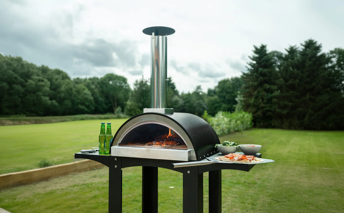Outdoor pizza oven with a sleek black design, stainless steel chimney, and fresh ingredients on a table in a lush green setting