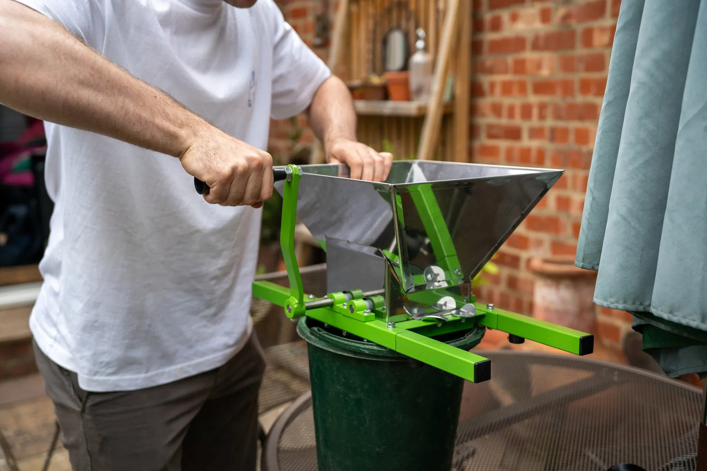 A person using a bright green fruit crusher with a metal hopper, positioned over a black bucket on a patio table, surrounded by brick walls