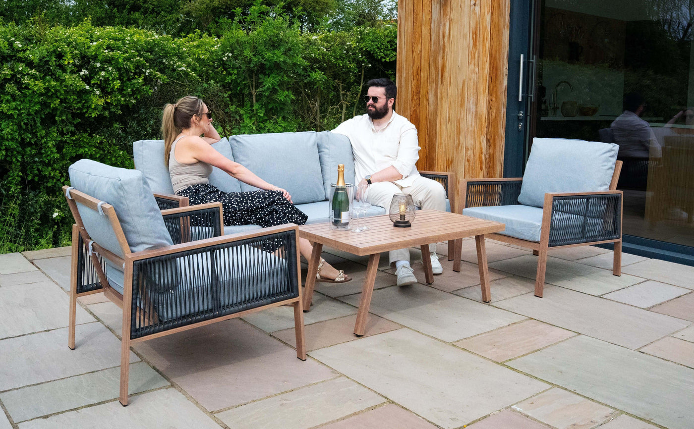 Stylish outdoor seating set featuring a wooden coffee table, light blue cushions, and a couple enjoying drinks in a garden setting