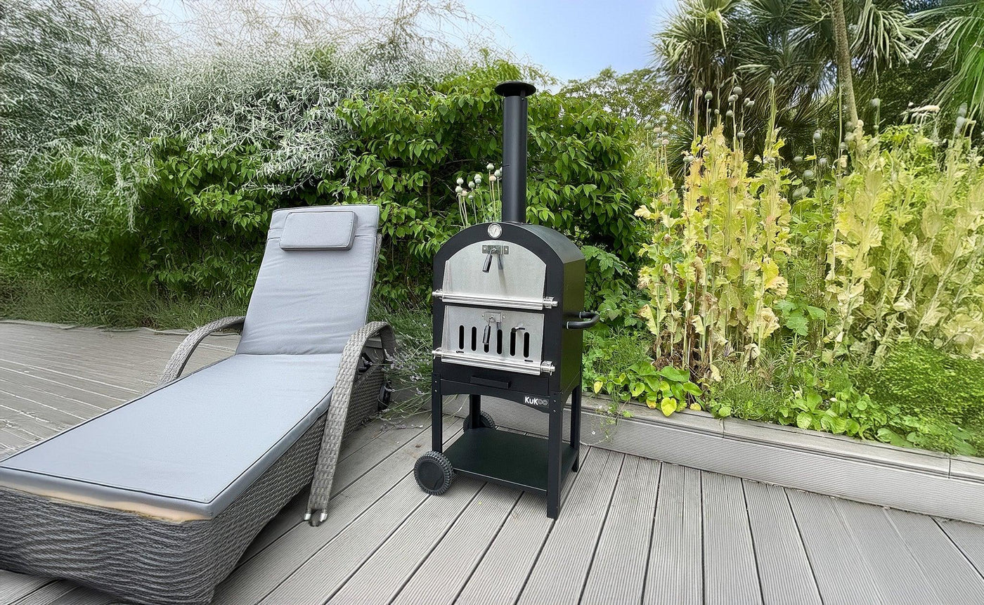 Black outdoor pizza oven on a wooden deck beside a gray lounge chair, surrounded by lush green plants and trees