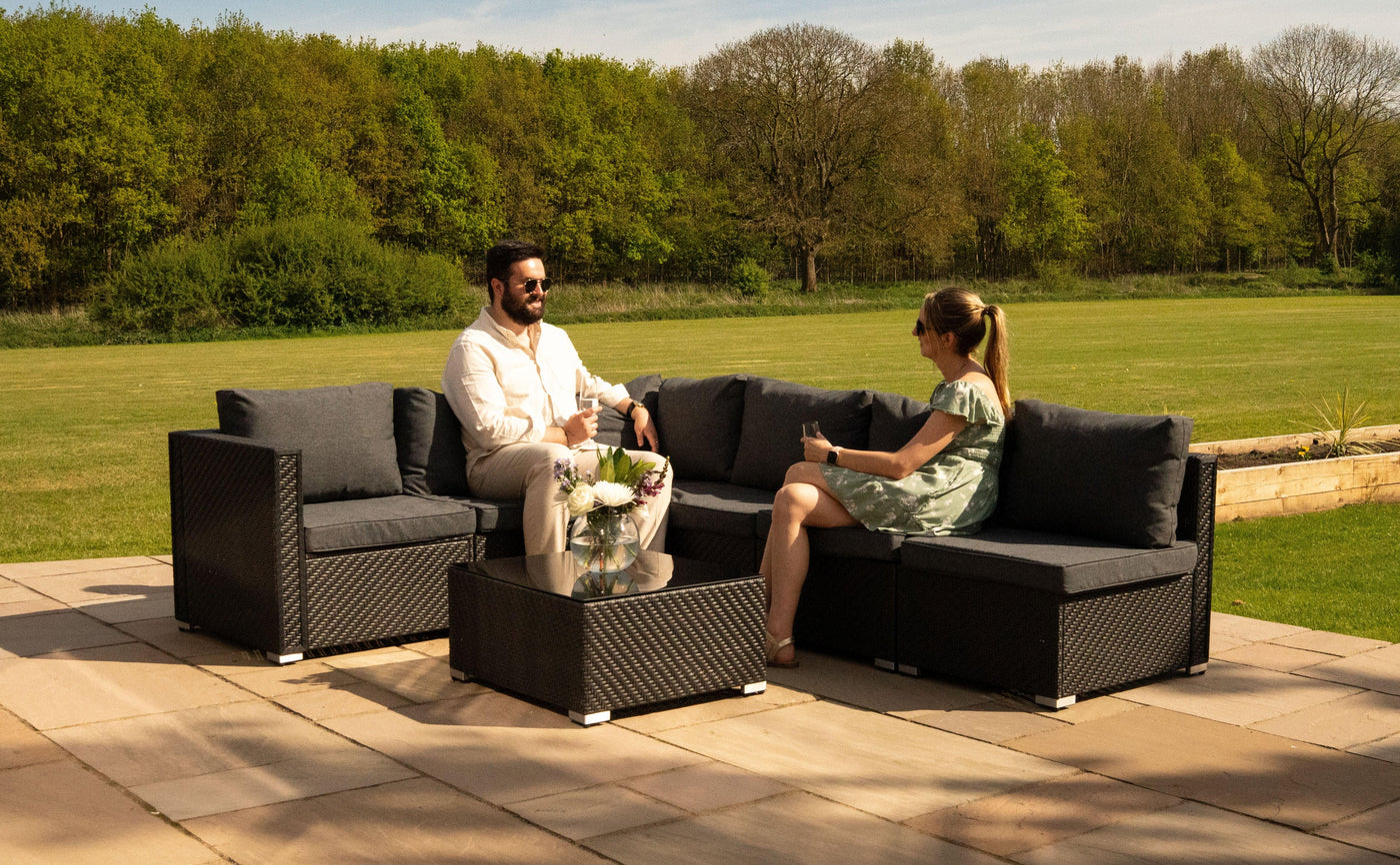 Stylish outdoor seating set featuring dark wicker furniture, gray cushions, and a glass-top coffee table, set in a lush green garden