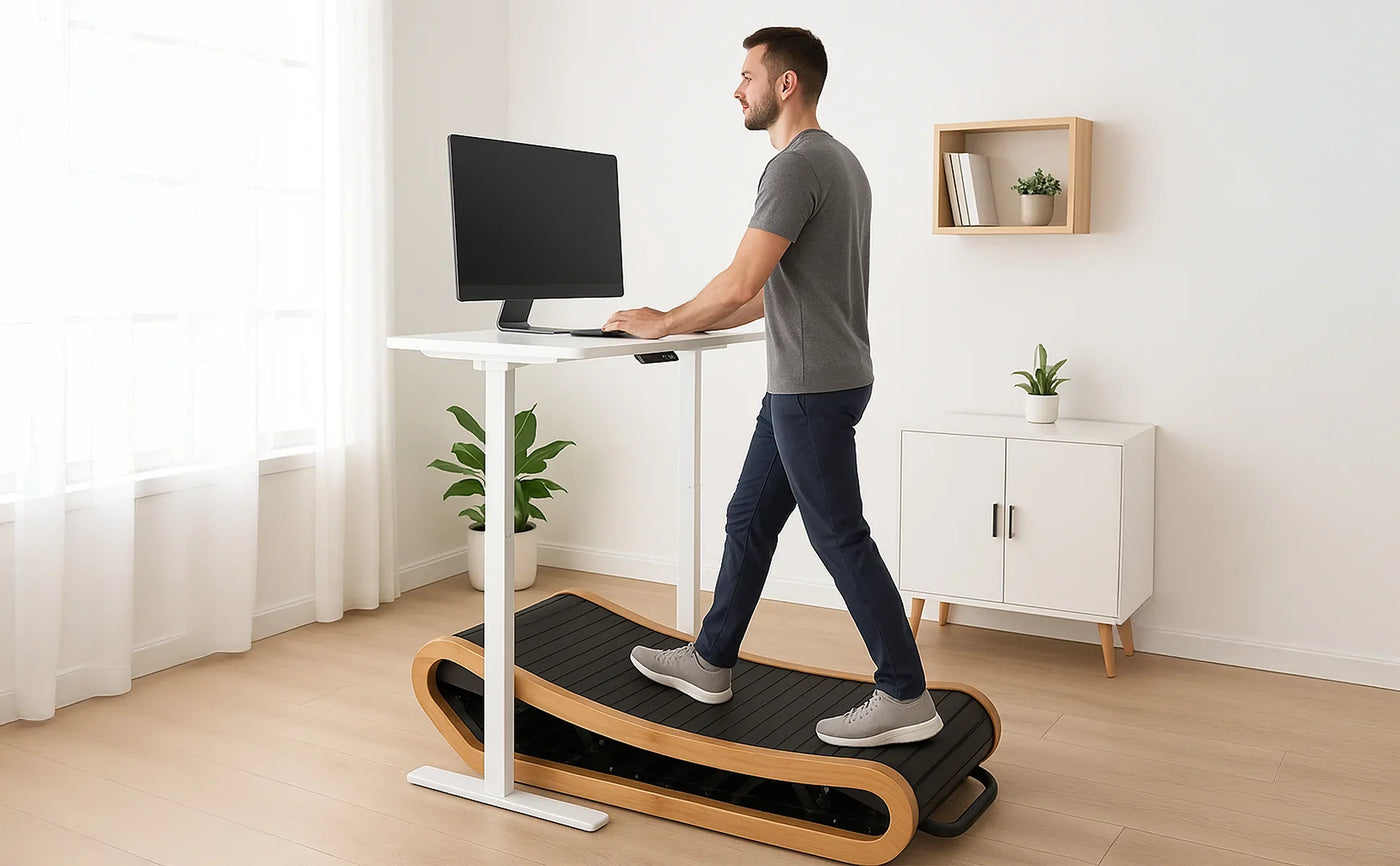 A man in a gray shirt walks on a sleek, wooden treadmill while working at a standing desk with a computer monitor in a bright room